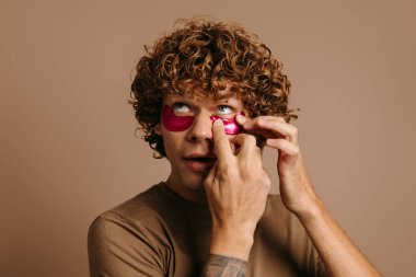 Yakışıklı kıvırcık adam kahverengi arka planda daha iyi cilt bakımı için yüz maskesi uyguluyor.