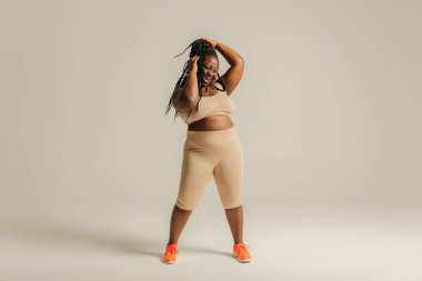 Spor giyimli Afrikalı güzel ve cüsseli bir kadın stüdyo arka planında rastalarını ayarlıyor.