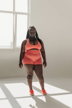 Spor giyim bölümünde kendinden emin, iri yarı Afrikalı bir kadın fitness stüdyosunda duruyor.