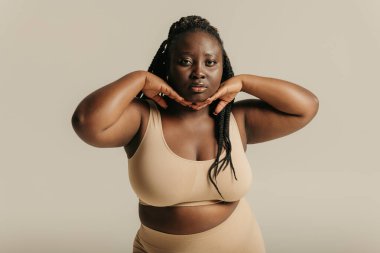 Güzel kıvrımlı Afrikalı kadın iç çamaşırlarıyla yüzüne dokunuyor ve stüdyo arka planındaki kameraya bakıyor.