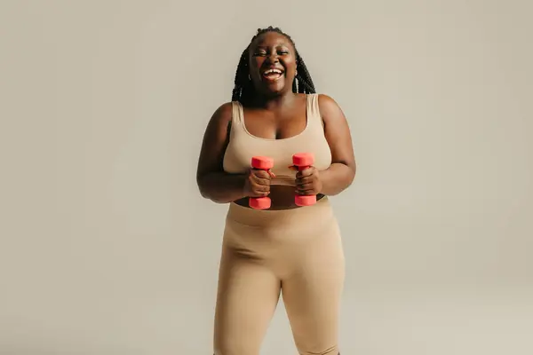 穿着运动服 带哑铃 带工作室背景的快乐加体形的非洲女人 — 图库照片