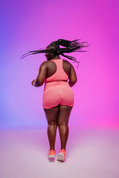 stock image Rear view of African curvy woman in sportswear dancing and looking confident against vibrant background