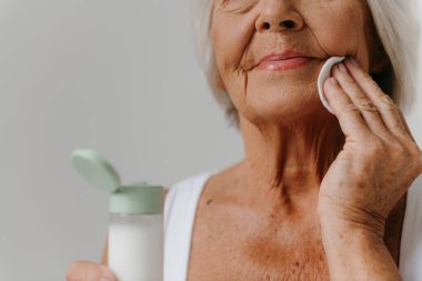 Gri arka planda pamuklu süngerle yüzünü temizleyen son sınıf kadınının yakın çekimi