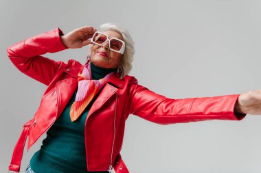 Gri arka planda dans ederken moda gözlüklü, deri ceketli, modaya uygun yaşlı bir kadın.