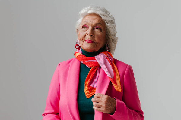 Elegant senior woman with make-up wearing fashionable clothing and radiating confidence on grey background