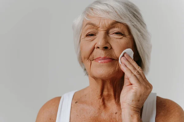 Confident senior woman cleaning face with cotton pad and looking at camera against grey background