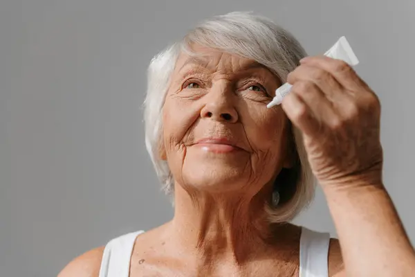 Confident senior woman applying anti-aging cream on face against grey background