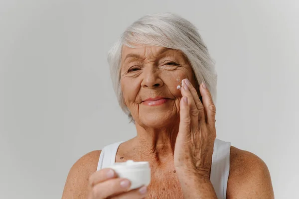 Confident senior woman applying anti-aging cream on face against grey background