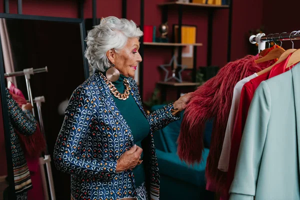 Moda mağazasında askıda asılı kıyafetleri inceleyen son sınıf öğrencisi, gri saçlı bir kadın.