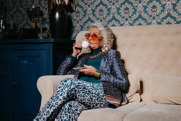 Elegant senior woman in trendy and luxury wear drinking coffee while sitting on the couch at retro styled home