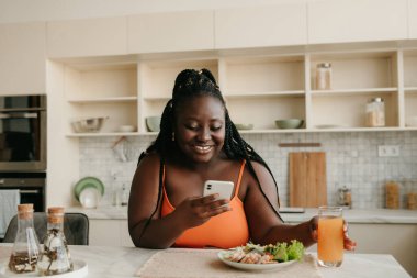 Güzel, iri yarı, akıllı telefon kullanan Afrikalı bir kadın evde öğle yemeği yerken sağlıklı beslenmenin tadını çıkarıyor.