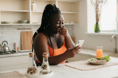 Neşeli ve iri Afrikalı kadın evde sağlıklı bir öğle yemeği yerken akıllı telefon kullanıyor.