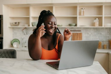 Mutlu ve iri Afrikalı kadın cep telefonuyla konuşuyor ve evde çalışırken dizüstü bilgisayar kullanıyor.