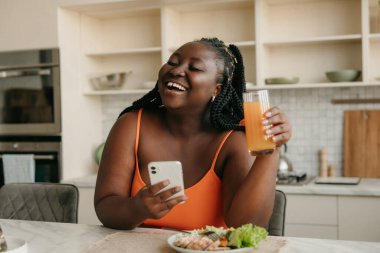 Neşeli ve cüsseli Afrikalı bir kadın akıllı telefon tutarken evde sağlıklı bir şekilde yemek yiyor.