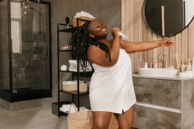 Banyoda şarkı söylerken kozmetik şişesini mikrofon gibi tutan neşeli, tombul Afrikalı kadın.