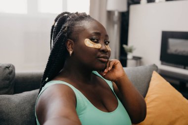 Güzel, iri yapılı, yüzünde yamalar olan, evde spa gününün keyfini çıkarırken selfie çeken Afrikalı bir kadın.