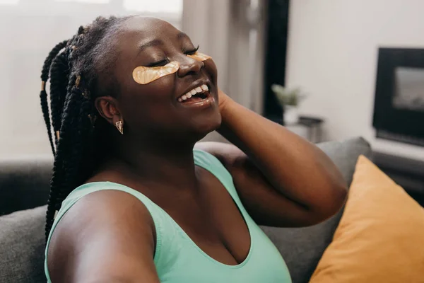 Joyful curvy African woman with eye patches on face making selfie while enjoying spa day at home