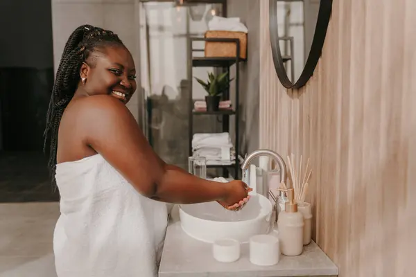 Happy curvy African woman covered in towel washing hands at the domestic bathroom