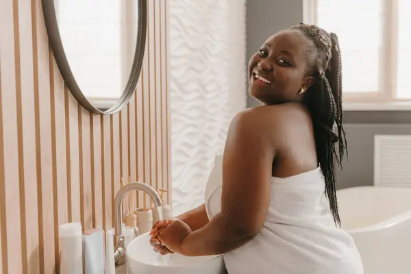 Joyful plus size African woman covered in towel washing hands and smiling at the domestic bathroom