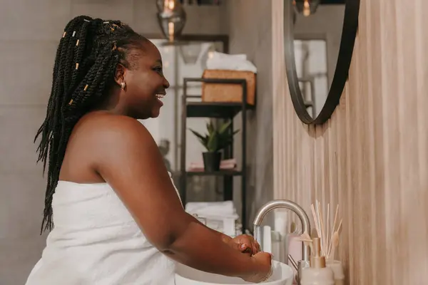 Beautiful plus size African woman covered in towel washing hands and smiling at the domestic bathroom