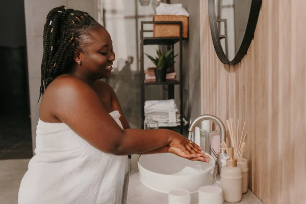 Happy plus size African woman covered in towel washing hands at the domestic bathroom