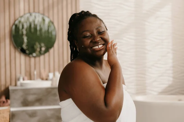 Beautiful curvy African woman covered in towel enjoying her smooth skin on face in bathroom