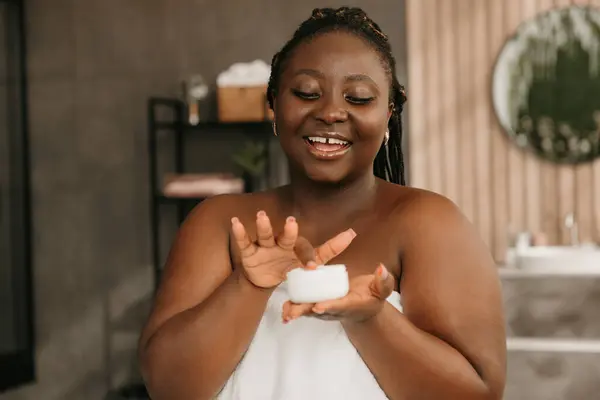 Beautiful plus size African woman covered in towel holding container with face cream in bathroom