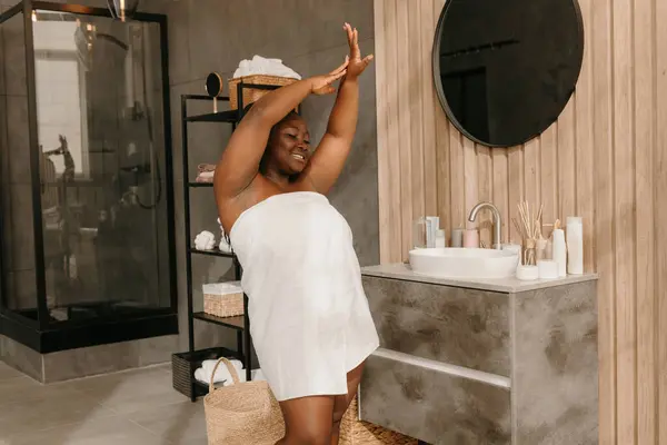 Happy plus size African woman covered in towel dancing in domestic bathroom