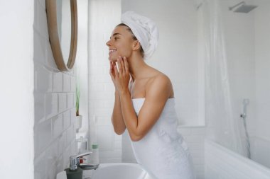 Havluya sarılı mutlu genç bir kadın banyoda dururken temiz ve taze yüzüne dokunuyor.