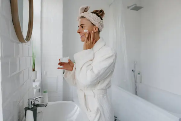 Beautiful young woman in bathrobe applying face cream and smiling while standing in bathroom