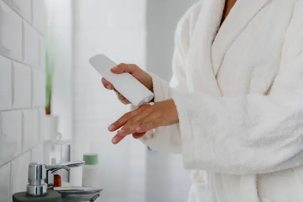 Close-up of woman in bathrobe applying cosmetic cream on hand while standing in bathroom