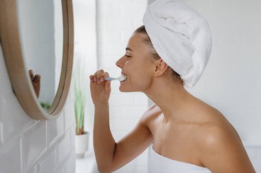 Kafasında havluyla banyoda dişlerini fırçalayan güzel genç bir kadın.