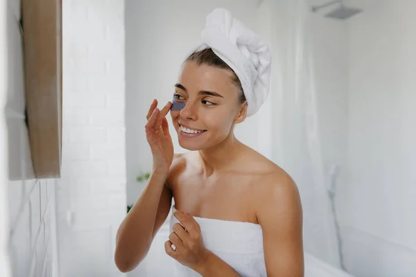 Attractive young woman covered in towel applying eye patches and smiling while standing in bathroom
