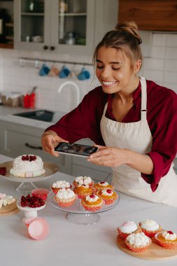 Mutfak masasında yatan çeşitli tatlıları çekerken akıllı telefon kullanan neşeli kadın fırıncı.