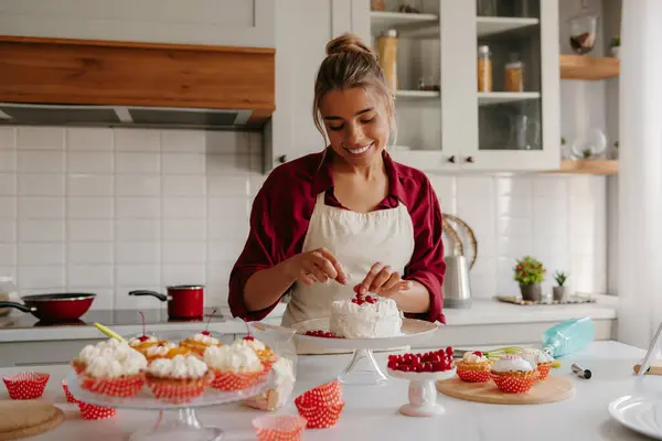 Beautiful female confectioner decorating cake with berries while standing at the domestic kitchen