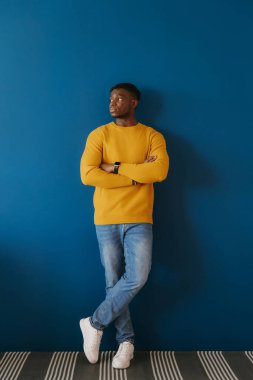 Kendine güvenen genç Afrikalı adam mavi duvara yaslanırken kollarını çapraz tutuyor.