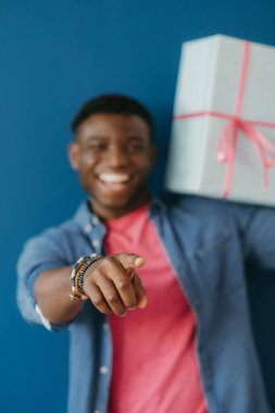 Sıradan giyinmiş, omuzunda hediye kutusu taşıyan ve mavi arka planda kamerası olan mutlu bir Afrikalı genç adam.