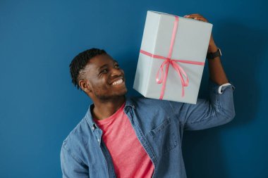 Sıradan giyinen yakışıklı Afrikalı bir adam hediye kutusunu omuzunda taşıyor ve mavi arka planda gülümsüyor.