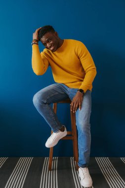 Mavi duvara karşı sandalyede otururken rahat görünen, gündelik giysili yakışıklı genç Afrikalı adam.