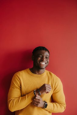 Kırmızı arka planda dikilirken kameraya gülümseyen gündelik giysili yakışıklı Afrikalı genç adamın portresi.