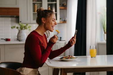 Genç ve mutlu bir kadının ev mutfağında sağlıklı bir öğle yemeğinin tadını çıkarırken akıllı telefon kullanması.