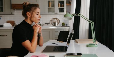 Yoğunlaşmış genç bir kadın masa başında oturmuş evde çalışırken dizüstü bilgisayara bakıyor.