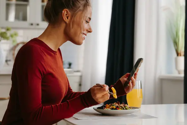 Smiling young woman using smart phone while enjoying healthy lunch at the domestic kitchen