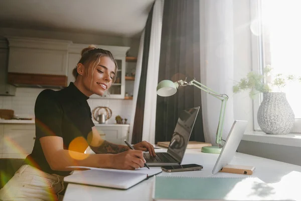 Confident young woman making notes in her note pad and smiling while working at home