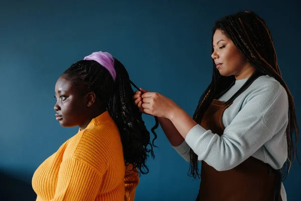 Side view of confident African hairdresser braiding hair to a female customer on blue background