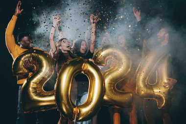 2024 numara tutan ve gece kulübünde yeni yılı kutlarken konfeti atan bir grup neşeli insan.