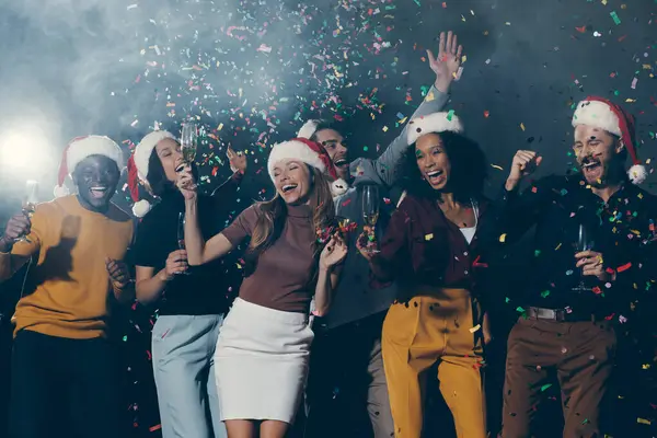 Joyful young people in Christmas hats dancing and throwing confetti while celebrating New Year in night club