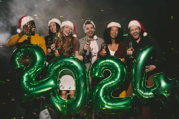Group of joyful people in Christmas hats holding 2024 numbers while celebrating New Year in night club