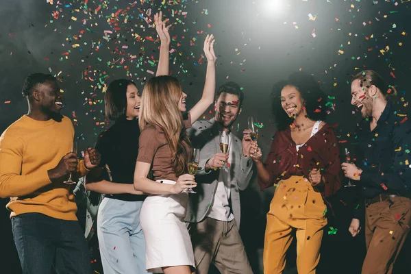 Group of young joyful people enjoying champagne and throwing confetti while dancing in night club together
