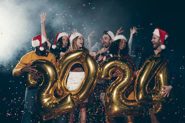 Group of happy people in Christmas hats celebrating 2024 New Year while throwing confetti in night club